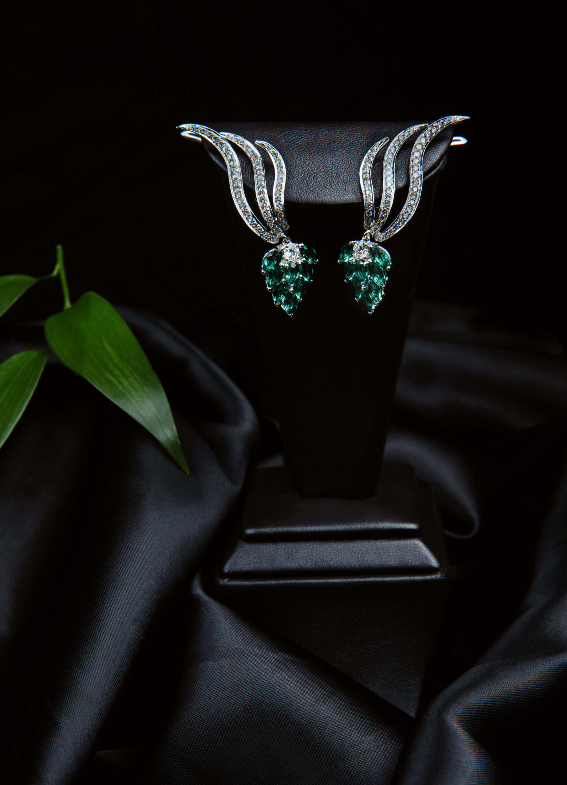 side view of a pair of silver diamond earrings with emerald on black background on black